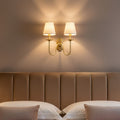 Clérachic twin-arm brass wall sconce lit above a channel-tufted taupe headboard, warm light fanning up the wall in soft rays.