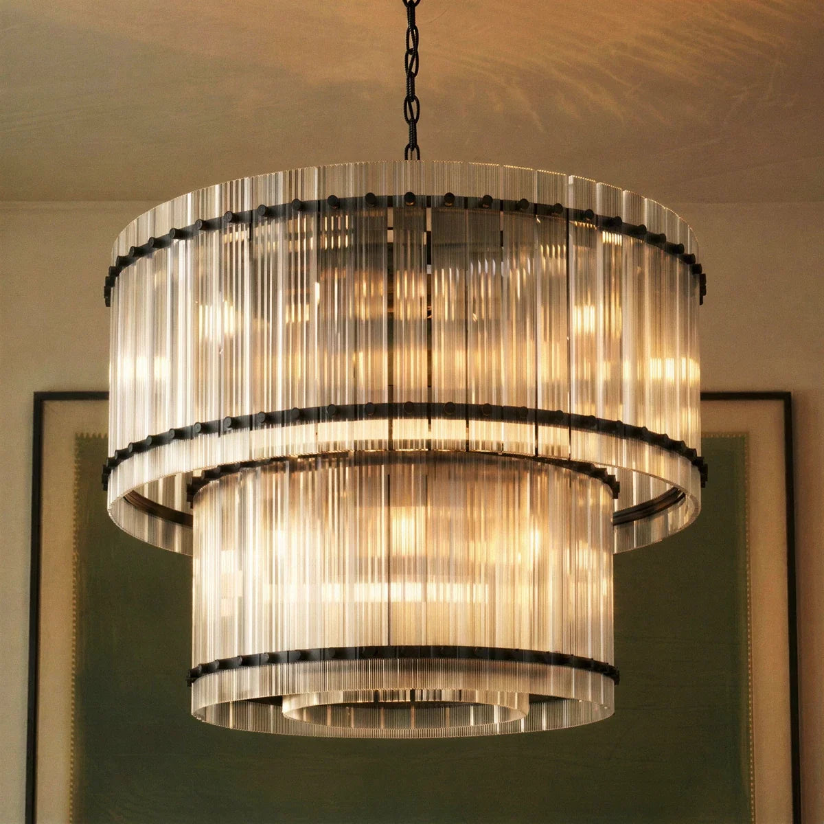 Clérachic close view of the matte black two-tier chandelier, with fluted glass rods lit warmly in front of a framed print.