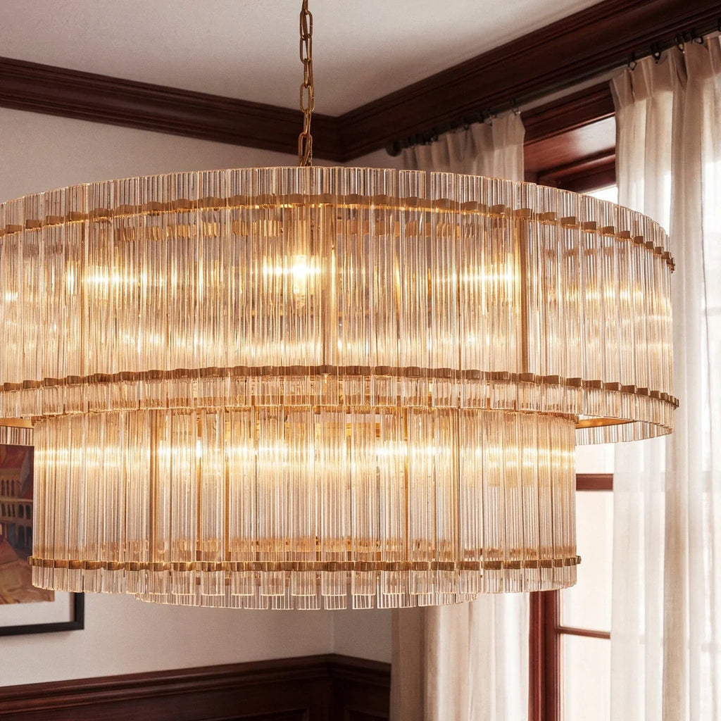 Clérachic close view of the antiqued brass chandelier, with fluted glass rods and brass bands beside a window with sheer curtains.