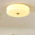 Warmly lit Clérachic two-tier flush mount ceiling light in cream glass with a brass finial, greenery blurred below.