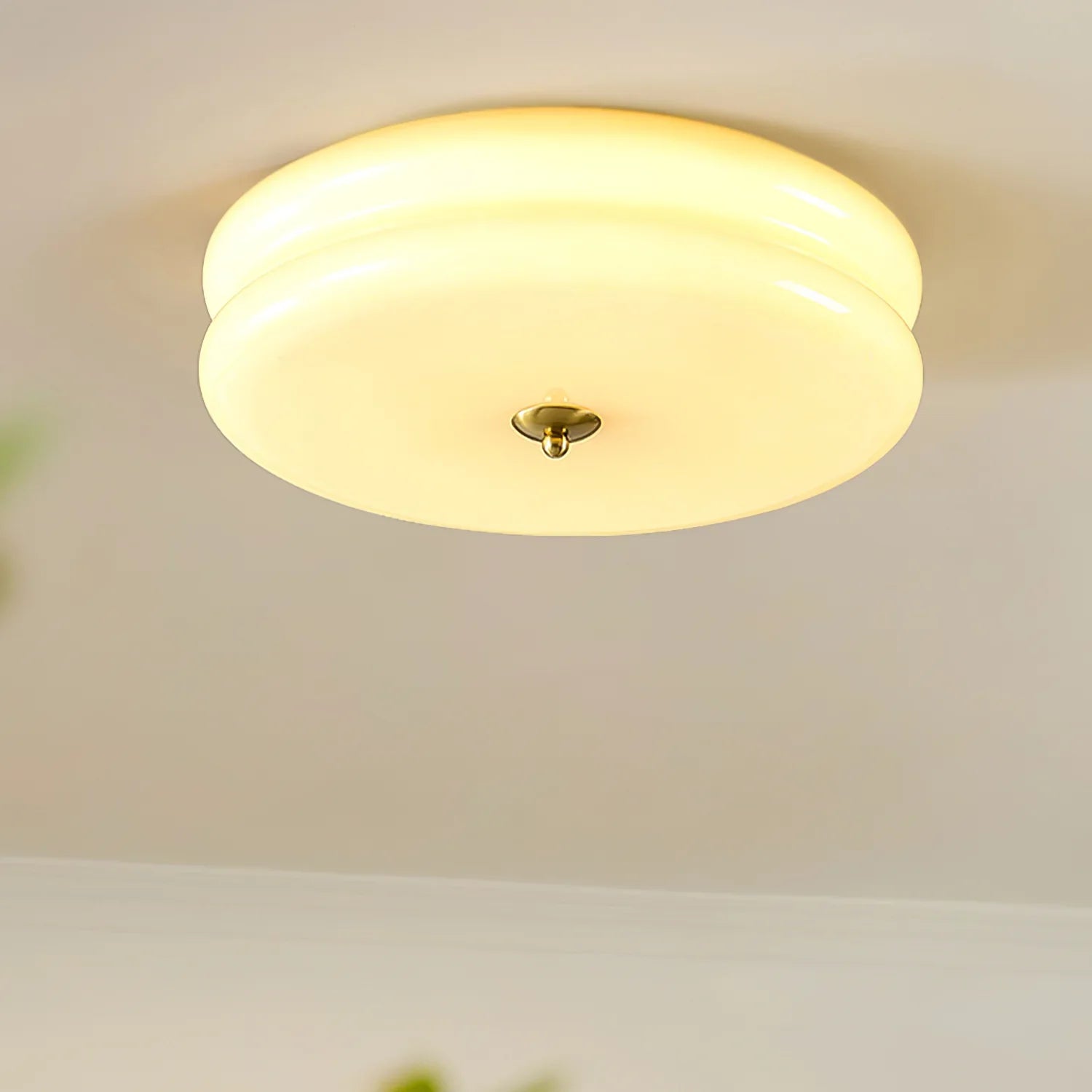 Warmly lit Clérachic two-tier flush mount ceiling light in cream glass with a brass finial, greenery blurred below.