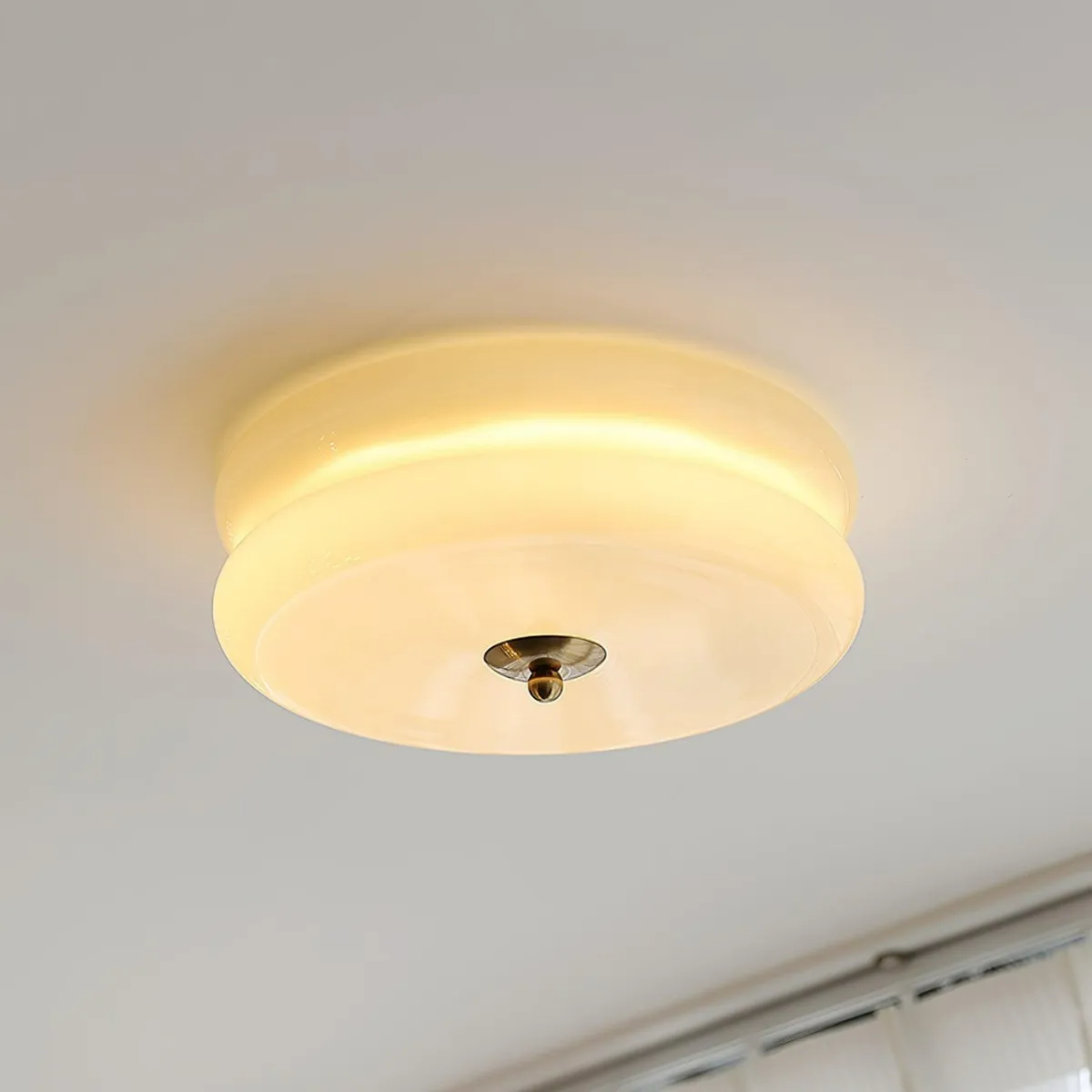 Clérachic two-tier flush mount ceiling light lit in cream glass, brass finial above a curtain rail and window.