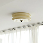 Unlit beige Clérachic two-tier flush mount ceiling light with a brass finial above a curtained window.
