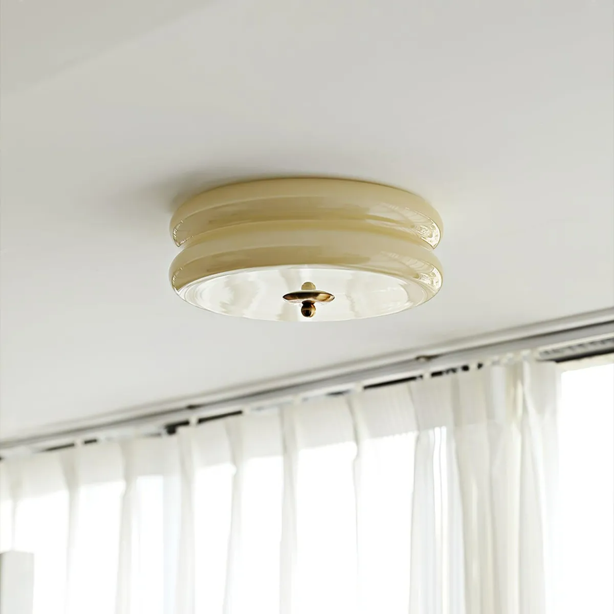 Unlit beige Clérachic two-tier flush mount ceiling light with a brass finial above a curtained window.