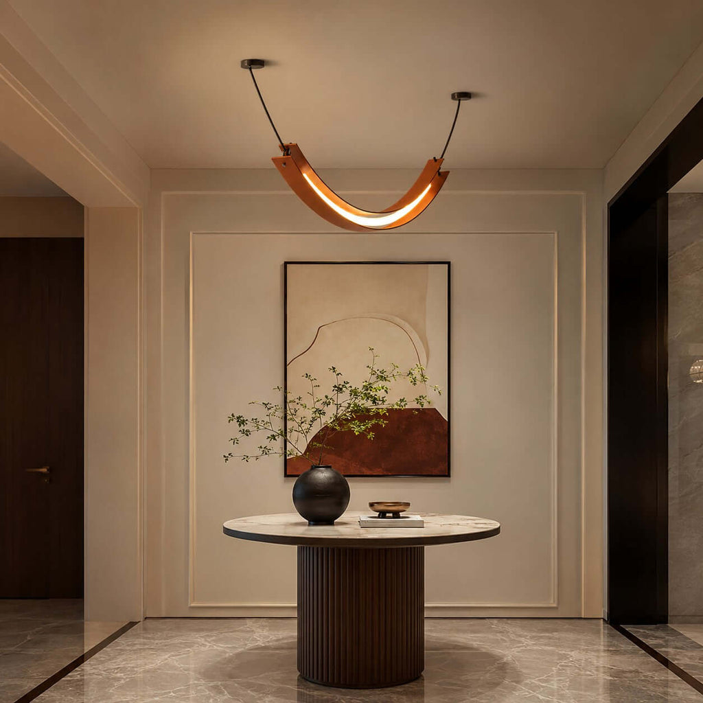 Lit terracotta U-shaped Clérachic pendant over a round fluted-base table in a hall, with a large abstract painting behind.