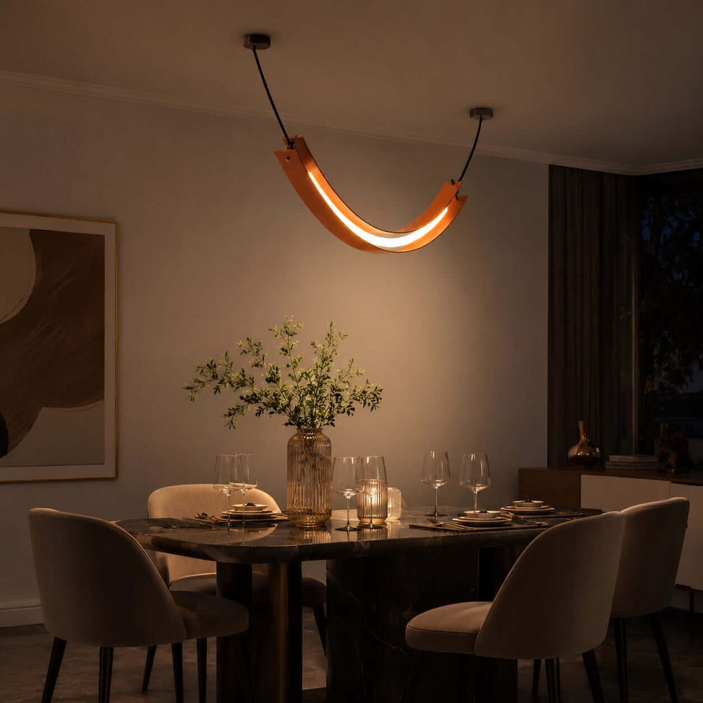A Clérachic U-shaped pendant glows amber over a set dining table with glasses and a vase of stems, in a dimly lit room.