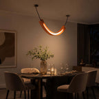 A Clérachic U-shaped pendant glows amber over a set dining table with glasses and a vase of stems, in a dimly lit room.