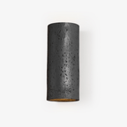 Black cylinder wall light with a pitted, stone-like surface and a warm gold glow inside the open end, on a pale backdrop; a Clérachic wall sconce.