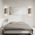 A pair of dark cylinder wall lights throw light up and down on either side of a framed artwork above a bed with grey linen; from Clérachic.