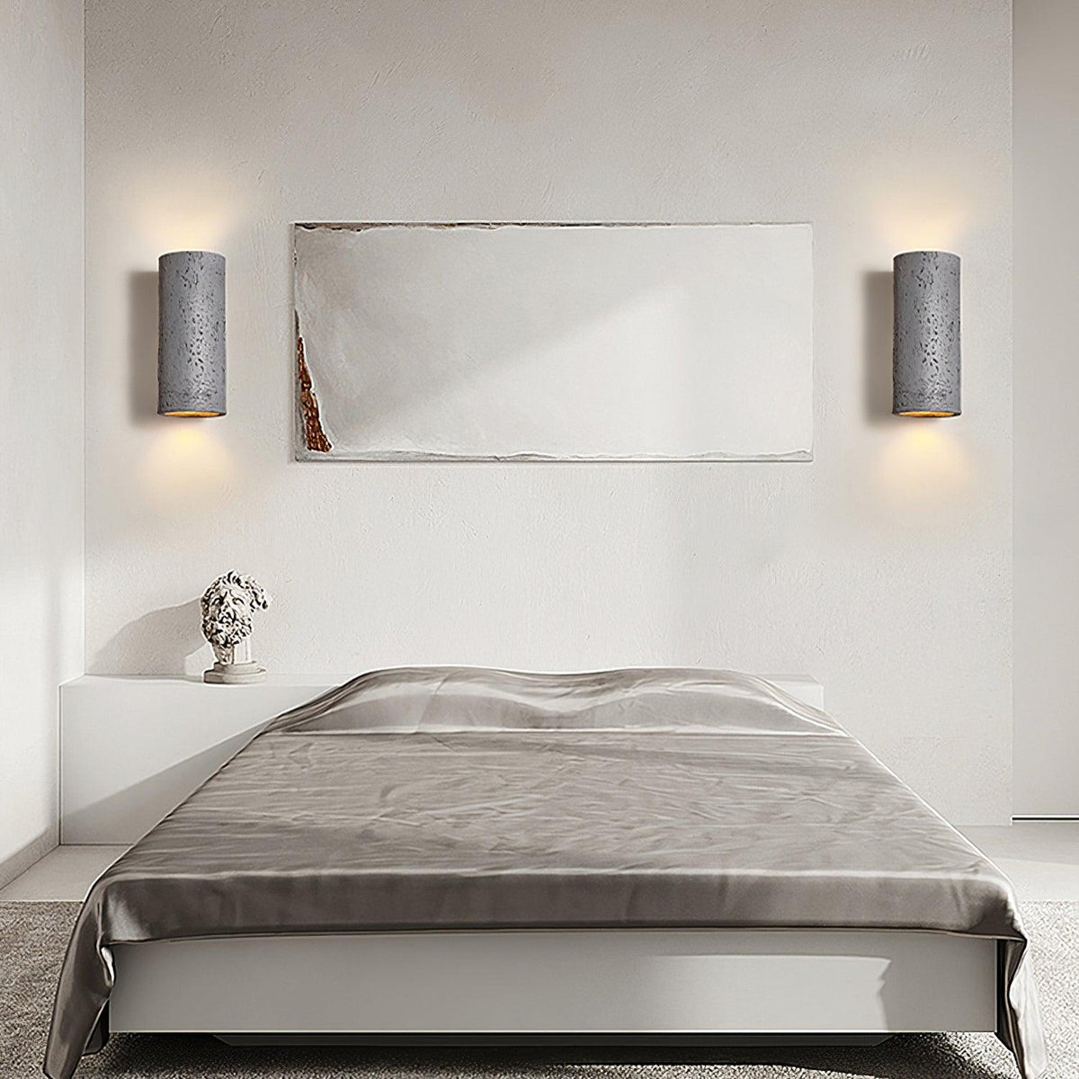 A pair of dark cylinder wall lights throw light up and down on either side of a framed artwork above a bed with grey linen; from Clérachic.