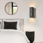 Black cylinder wall light glowing above a bed with white and charcoal pillows, beside a round mirror and a ribbed black side table; a Clérachic design.