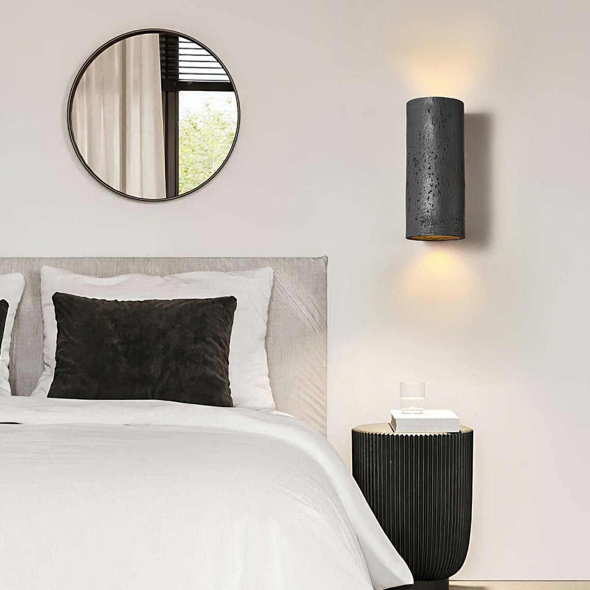 Black cylinder wall light glowing above a bed with white and charcoal pillows, beside a round mirror and a ribbed black side table; a Clérachic design.