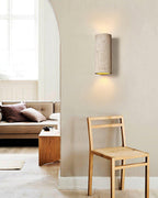 Pale travertine-look cylinder light on a curved wall beside a wooden chair and an arched opening, giving a soft wash; a Clérachic wall sconce.