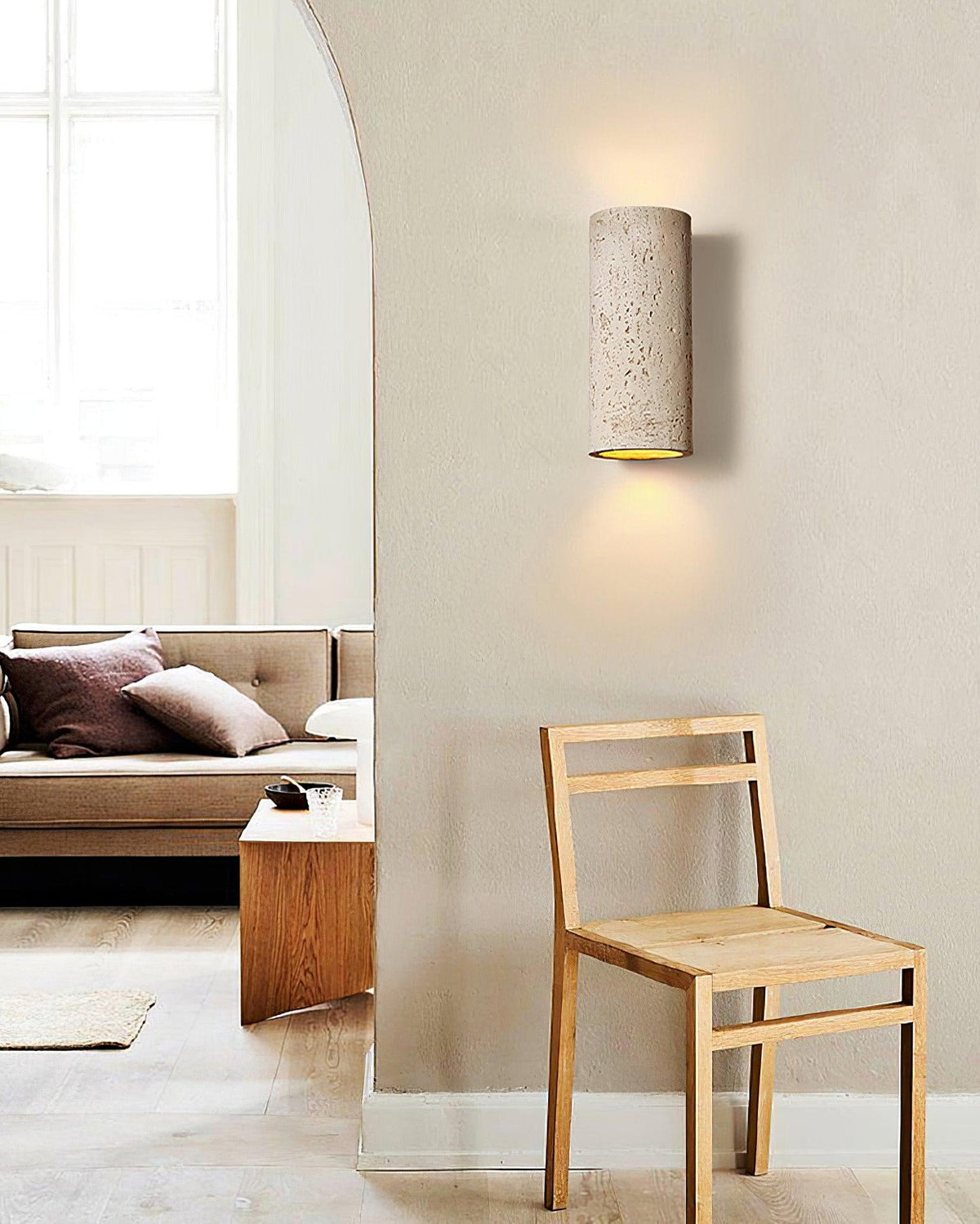 Pale travertine-look cylinder light on a curved wall beside a wooden chair and an arched opening, giving a soft wash; a Clérachic wall sconce.
