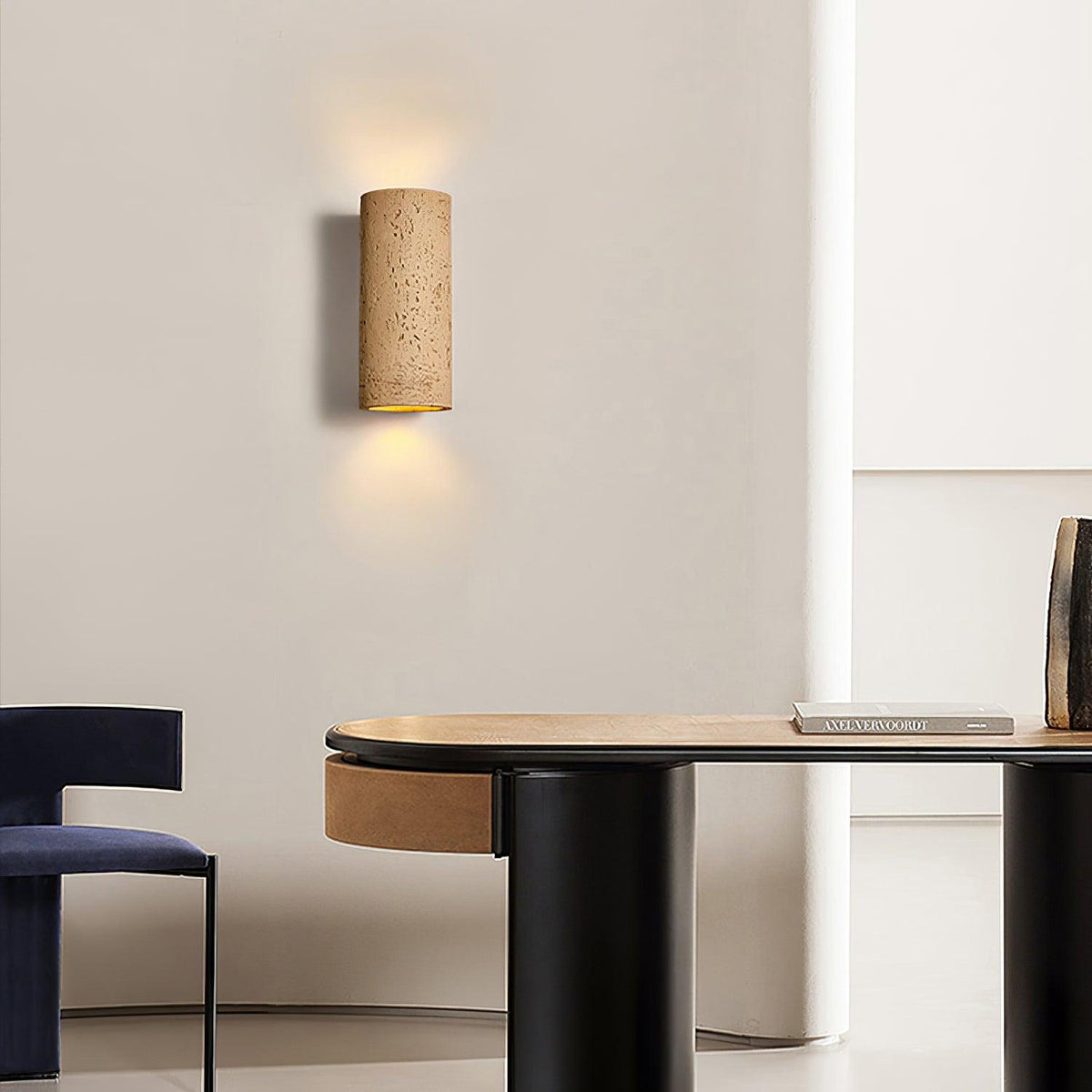 A tan cylinder wall light casts light up and down above a black-based desk and a navy chair; a Clérachic wall light.