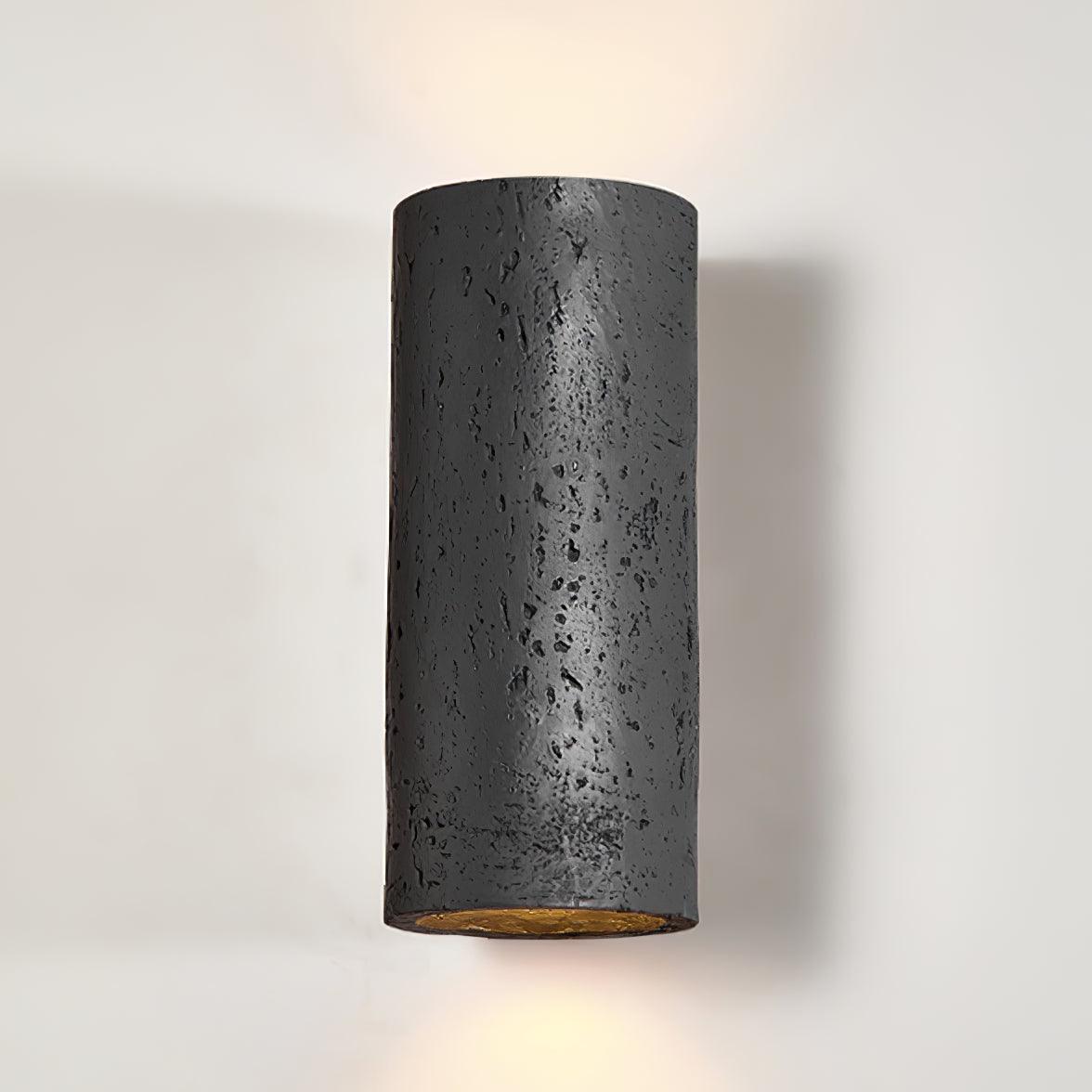 Black version of the cylinder light, close view, with light glowing from the bottom and a soft shadow on the wall; from Clérachic.