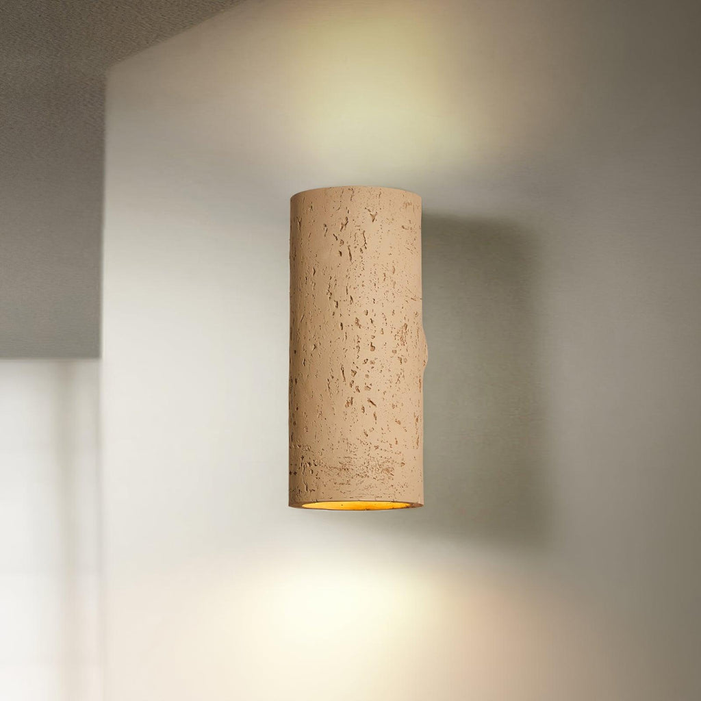 Tan cylinder wall light lit up and down, with a soft beam on the pale wall around it; a Clérachic wall sconce.