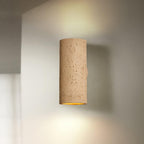 Tan cylinder wall light lit up and down, with a soft beam on the pale wall around it; a Clérachic wall sconce.