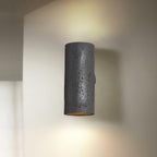 Black cylinder wall light with a pitted surface, casting a soft beam on the wall behind it; a Clérachic wall light.