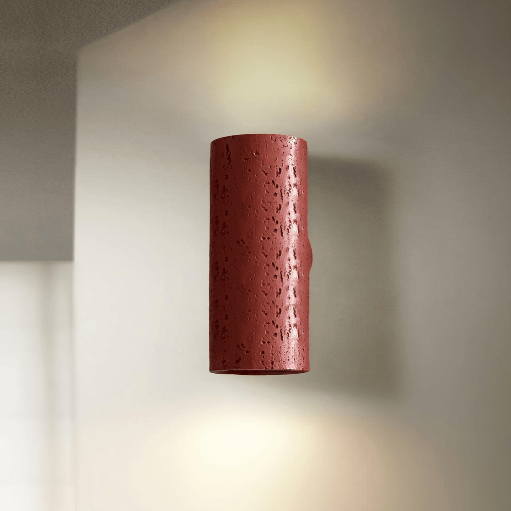A red cylinder wall light with a pitted finish glows up and down against a pale wall; a Clérachic wall sconce.