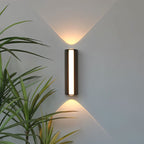 Clérachic slim black cylindrical wall lamp with a glowing front strip, casting warm up and down beams beside a palm on a grey wall.