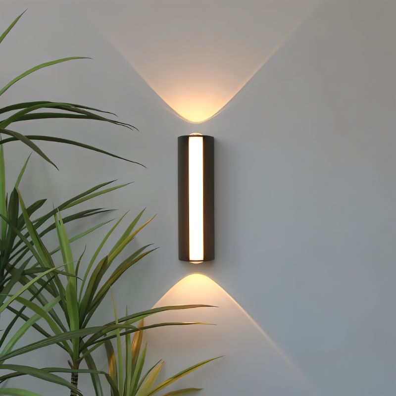 Clérachic slim black cylindrical wall lamp with a glowing front strip, casting warm up and down beams beside a palm on a grey wall.