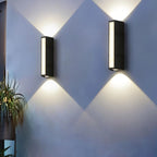 Clérachic pair of black rectangular wall lamps with side vents, casting crossing cool white beams on a pale grey wall.