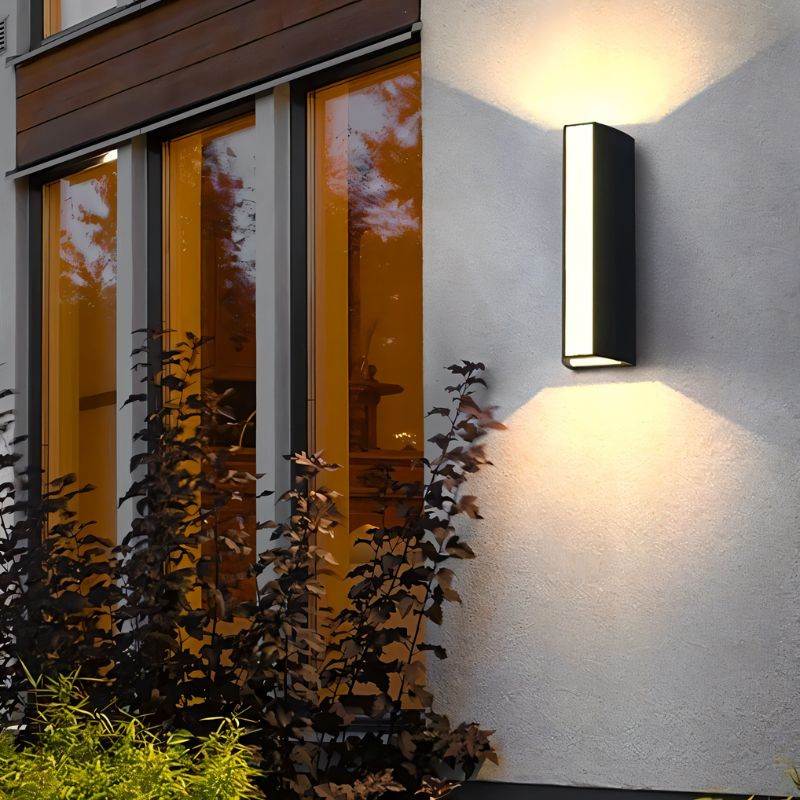 Clérachic matte black wall lamp on a rendered house wall at dusk, warm light fanning up and down beside lit glazed windows.