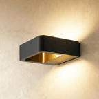 Clérachic square black aluminium wall light with a brass-toned inner reflector, throwing light up and down a plaster wall.