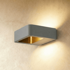 Clérachic square grey aluminium wall light with a brass-toned inner reflector, casting light up and down a plaster wall.
