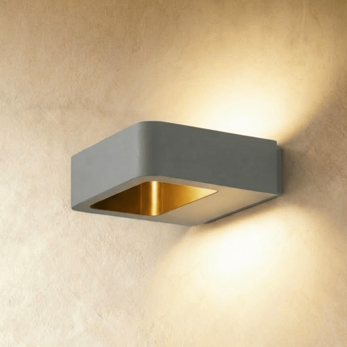 Clérachic square grey aluminium wall light with a brass-toned inner reflector, casting light up and down a plaster wall.