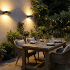 Two Clérachic black wall lights glowing at dusk on a courtyard wall above a laid timber table, with string lights in the greenery.
