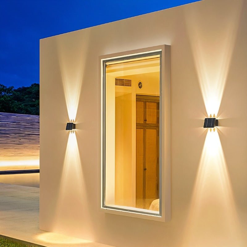 Two Clérachic solar wall lights throwing warm up-and-down beams on a pale exterior wall either side of a lit window at dusk.