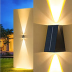 Clérachic black solar wall light in close-up, its up-and-down glow fanning across a pale outdoor wall above a lawn at dusk.