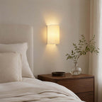 Clérachic fabric wall light glowing beside a cream headboard, above a dark wood bedside table with a vase of green sprigs.