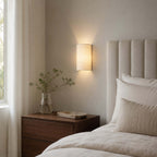 Clérachic half-round wall light glowing above a walnut bedside table, beside a ribbed headboard and a sheer curtain.
