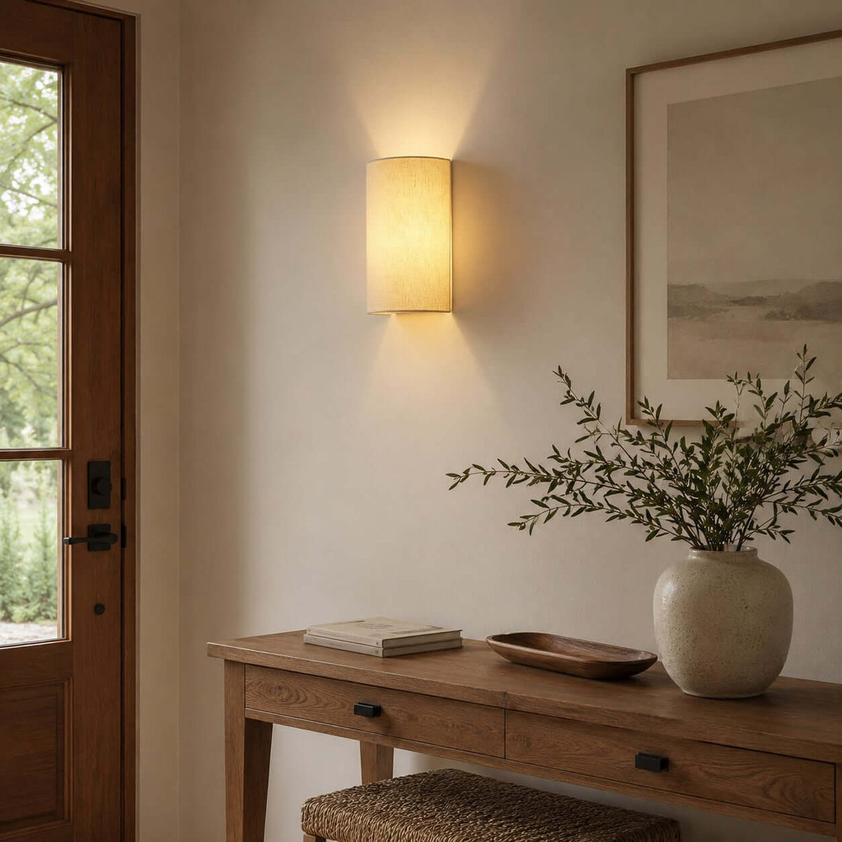 Clérachic wall light beside a wood-framed glass door, above a desk with a bowl, a vase of olive branches and a framed print.