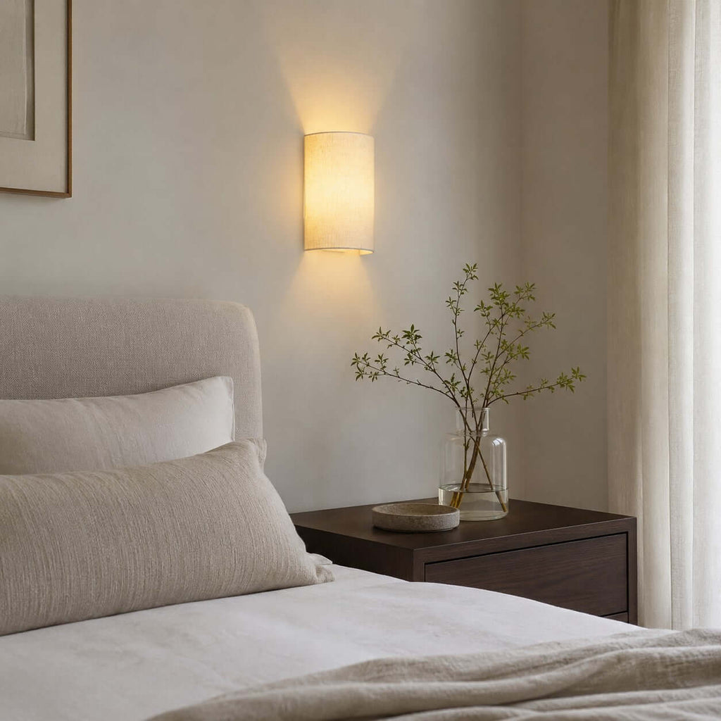 Clérachic half-round wall light above a walnut bedside table and a vase of spring stems, with a cream bed and a linen curtain.