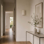 Unlit Clérachic half-round wall light in a bright hallway with a framed print, a metal console and a vase of olive branches.