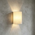 Close view of the linen-textured half-round shade on this Clérachic wall light, casting light up and down a grey plaster wall.