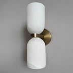 The Clérachic twin-cup alabaster sconce at an angle, polished brass band and round plate bright against a soft grey wall.