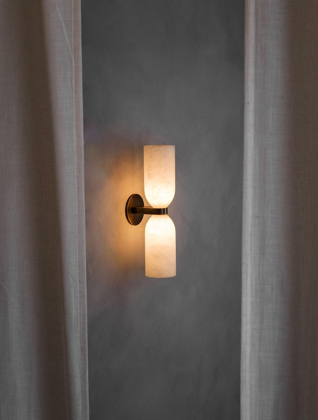 The Clérachic up-down sconce lit at dusk between linen curtains, washing the wall above and below with warm light.