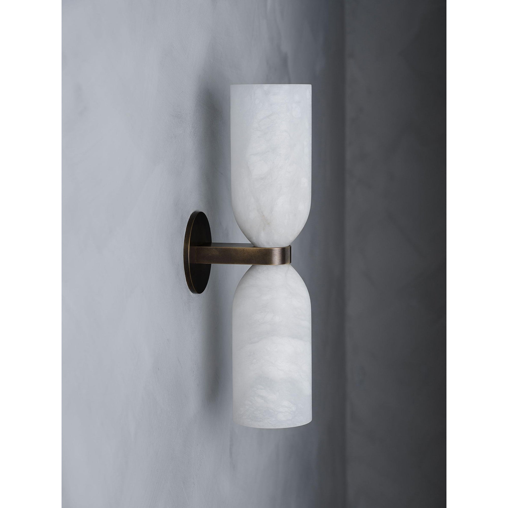 Angled view of the Clérachic wall sconce unlit, the aged brass arm and round plate reading warm against pale grey plaster.