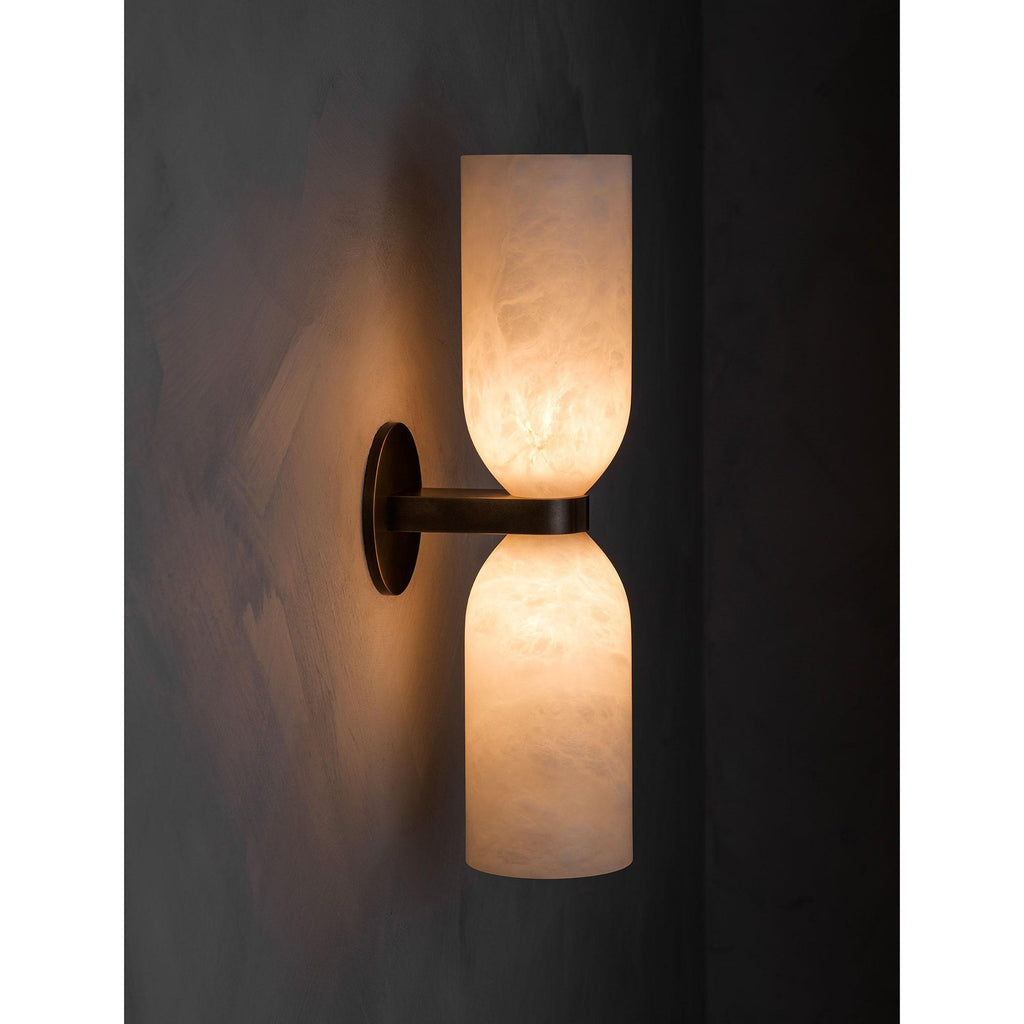 The Clérachic sconce lit in a darkened room, a soft halo of light behind the brass plate and no beam pointed at anything.