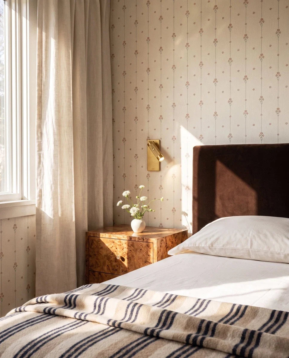 Clérachic brass wall reading light above a burr-wood bedside table, set against floral wallpaper beside a brown velvet headboard.