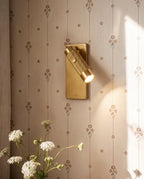 Clérachic brass reading spotlight on a rectangular plate, its knurled barrel angled down, on floral wallpaper beside wildflowers.