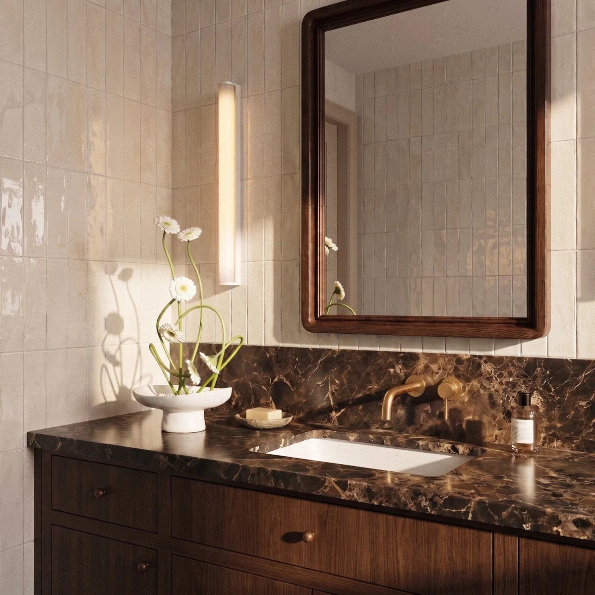 Clérachic slim vertical wall light glowing beside a walnut-framed mirror above a marble vanity and glazed cream tiles.