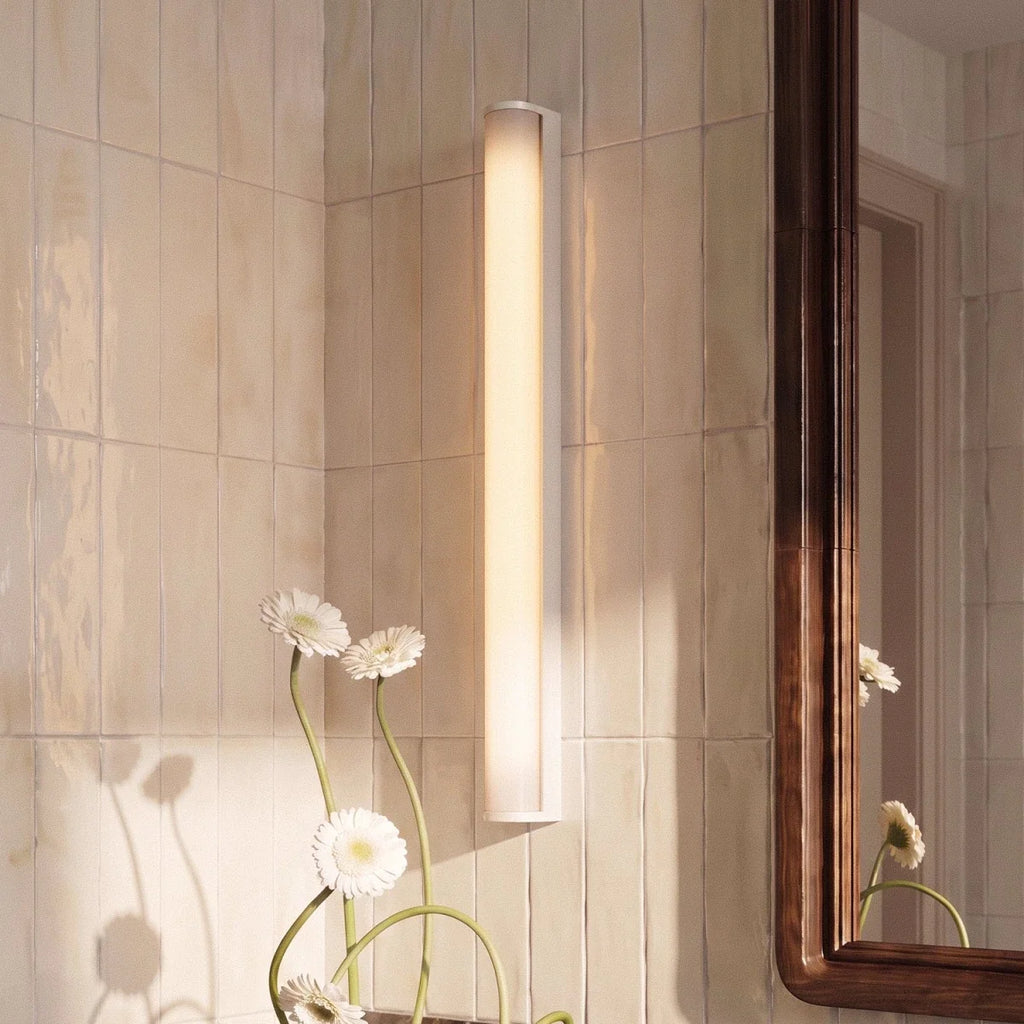 Clérachic white linear wall light mounted vertically on glossy cream tiles, with daisies and a walnut mirror frame beside it.