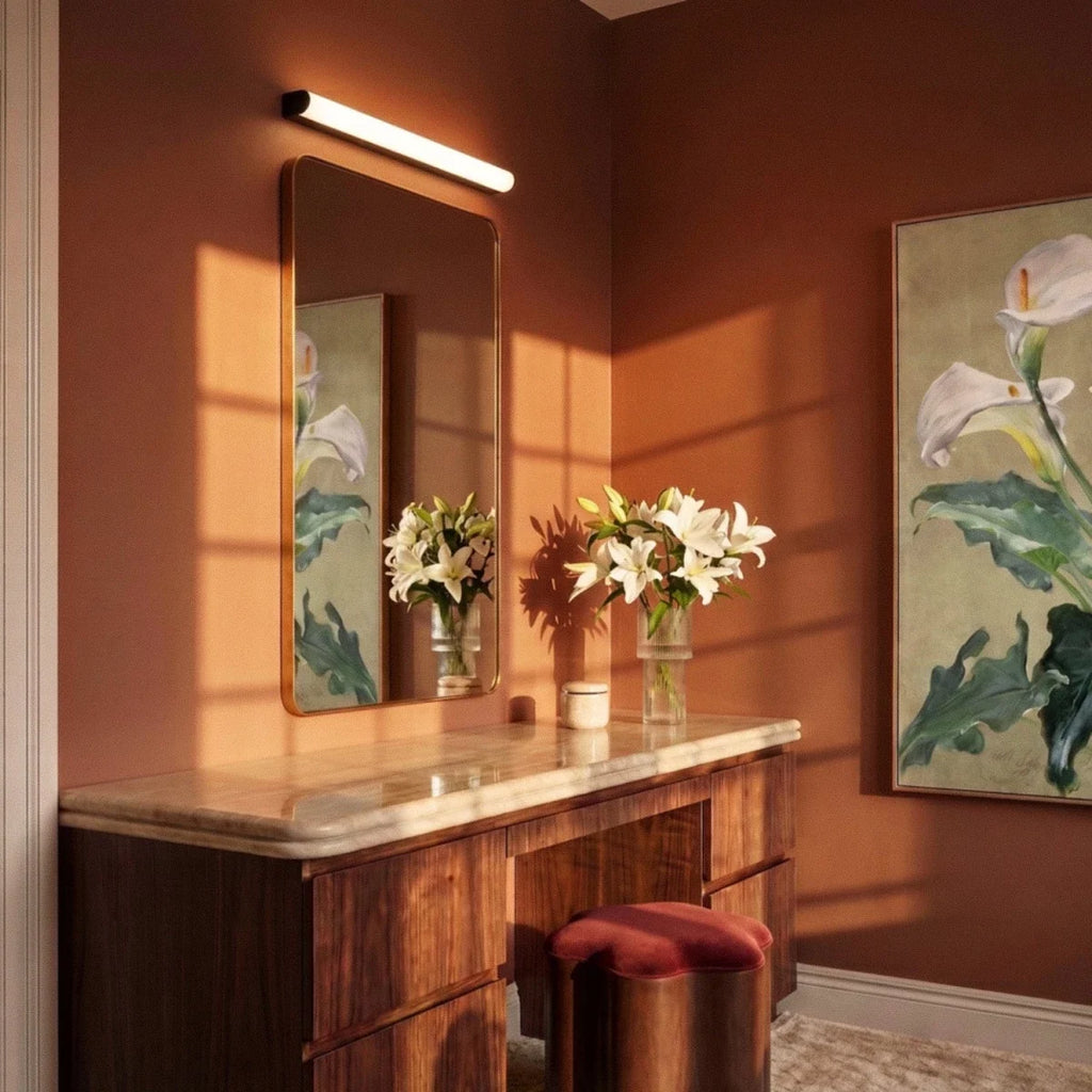 Clérachic linear wall light mounted above a gold-framed mirror at a walnut dressing table in a warm terracotta room.