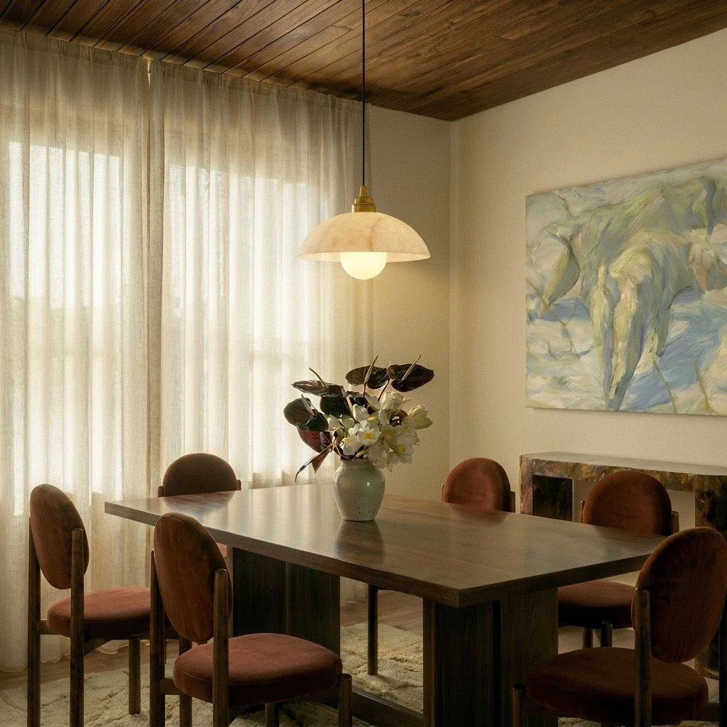 Clérachic alabaster dome pendant lit over a walnut dining table with velvet chairs, a vase of flowers and a large painting.